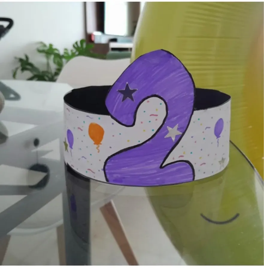 Corona de papel con un 2 morado, globos y estrellas, decorada por un niño